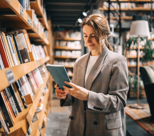 Young,Woman,Looking,At,Book,Taken,From,The,Shelf,In